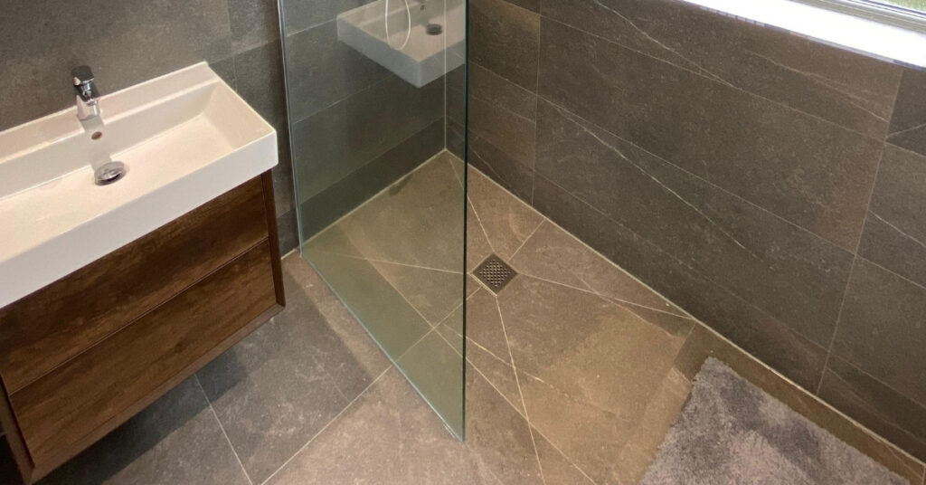 A modern accessible bathroom with a level‑access shower, glass screen, textured grey floor tiles, and a wooden vanity with a vessel basin.