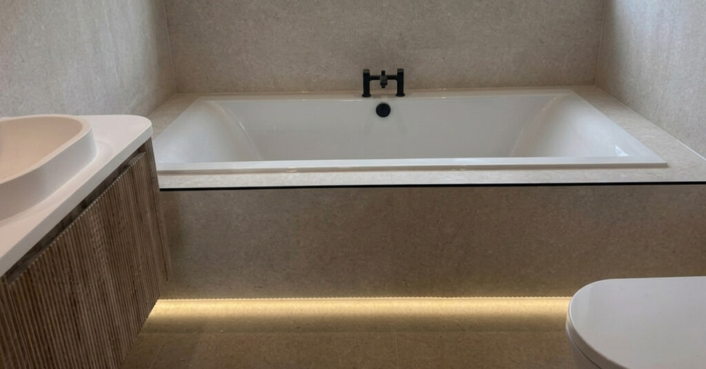 A contemporary spa‑style bathroom with a stone‑surround bathtub illuminated by soft LED under‑lighting, paired with a wooden vanity and vessel sink, all in warm neutral tones.
